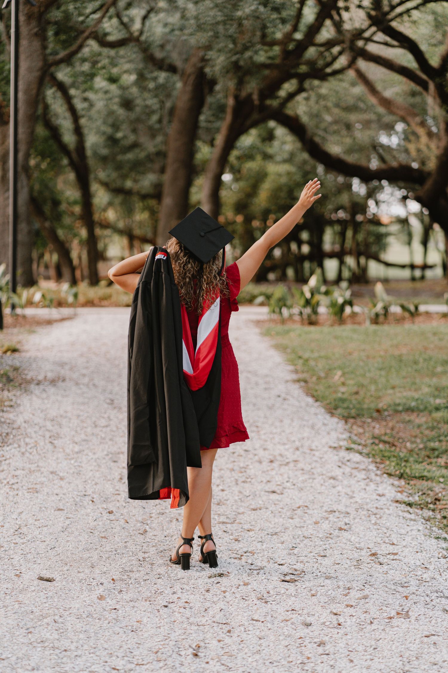 Graduate in red dress and black cap and gown celebrating on a gravel path surrounded by trees in a park setting.
