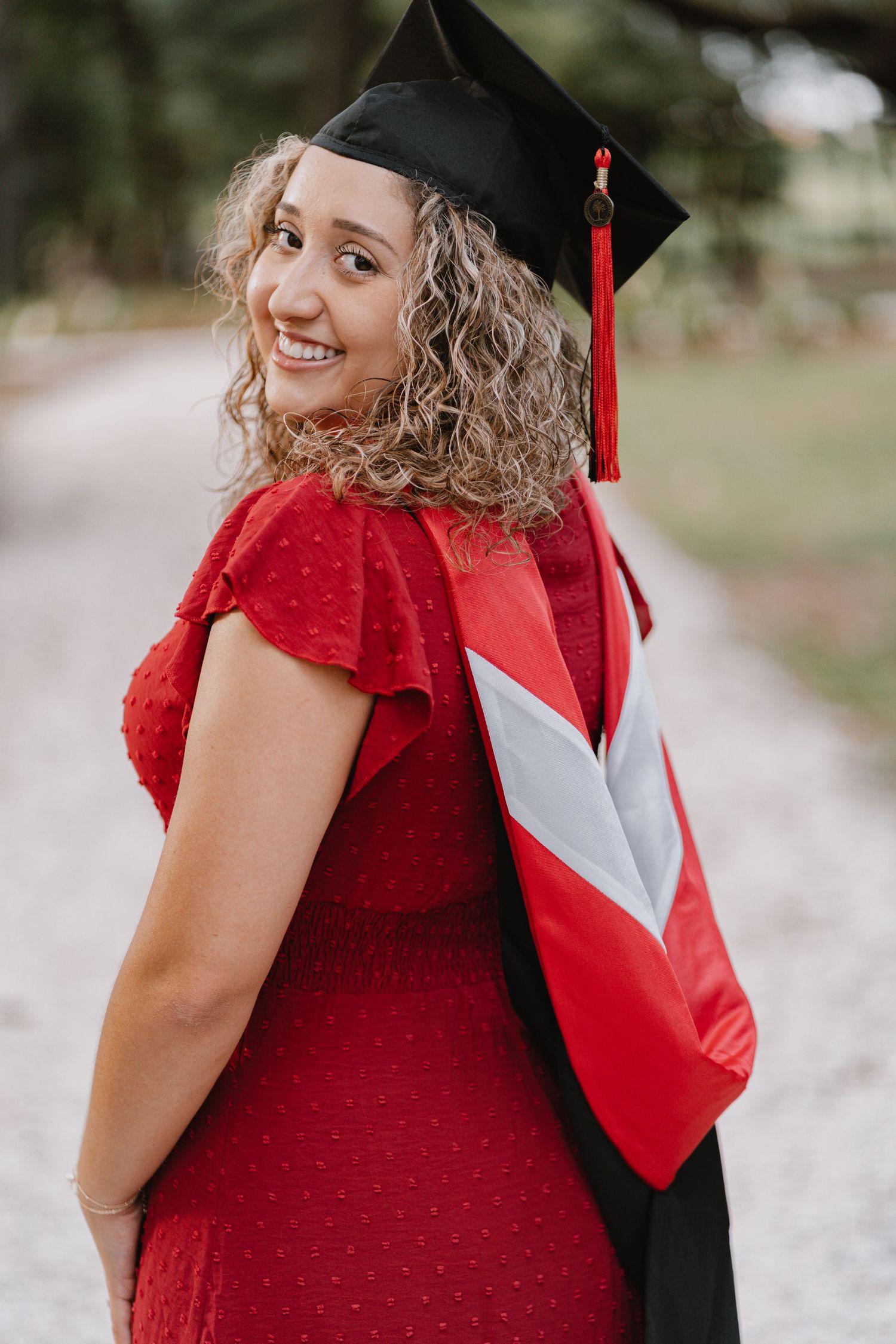 Graduate in red dress and cap with tassel looking over shoulder poses outdoors during graduation photo session.
