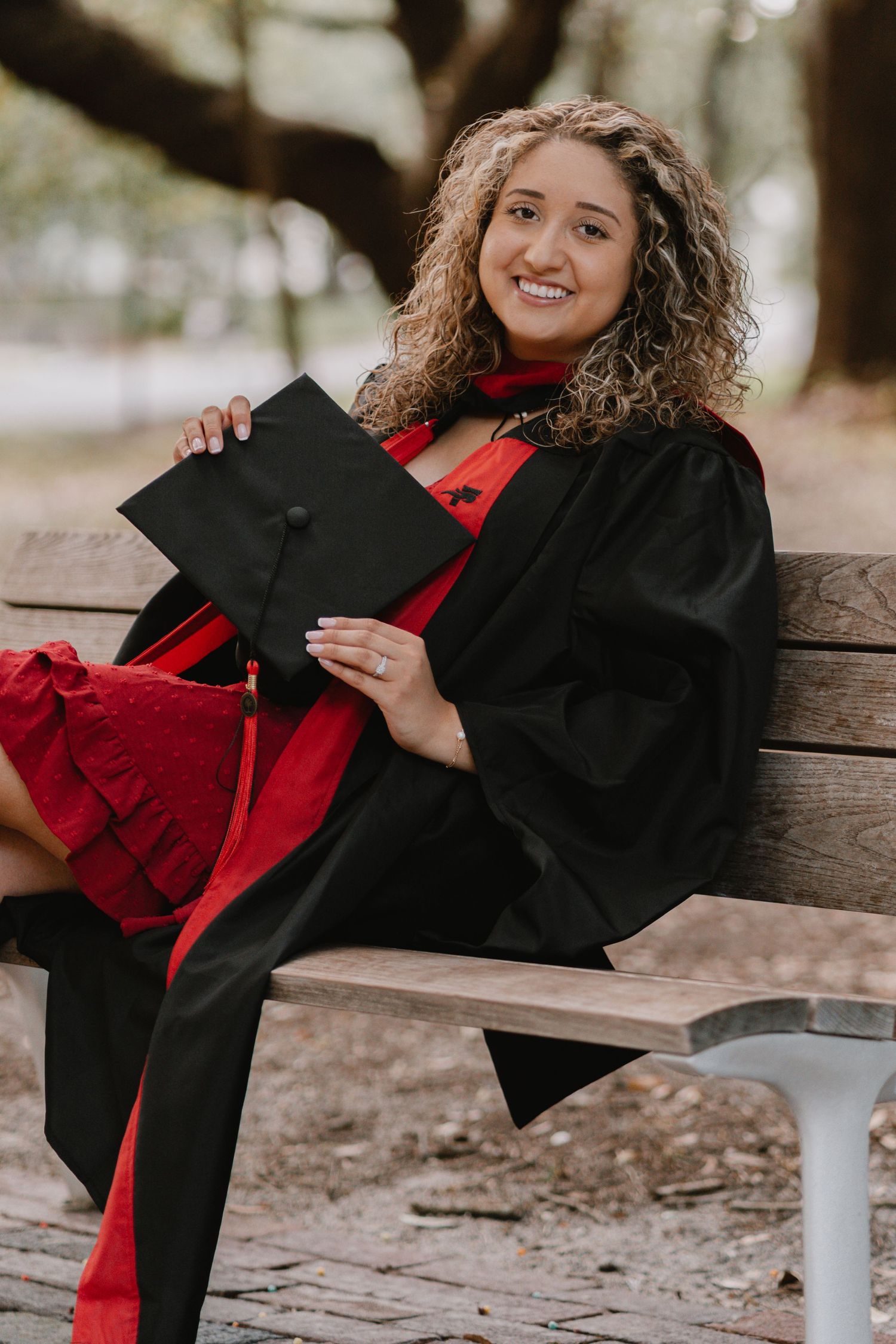 Graduate in black cap and gown with red dress sits on bench holding graduation cap while smiling outdoors.