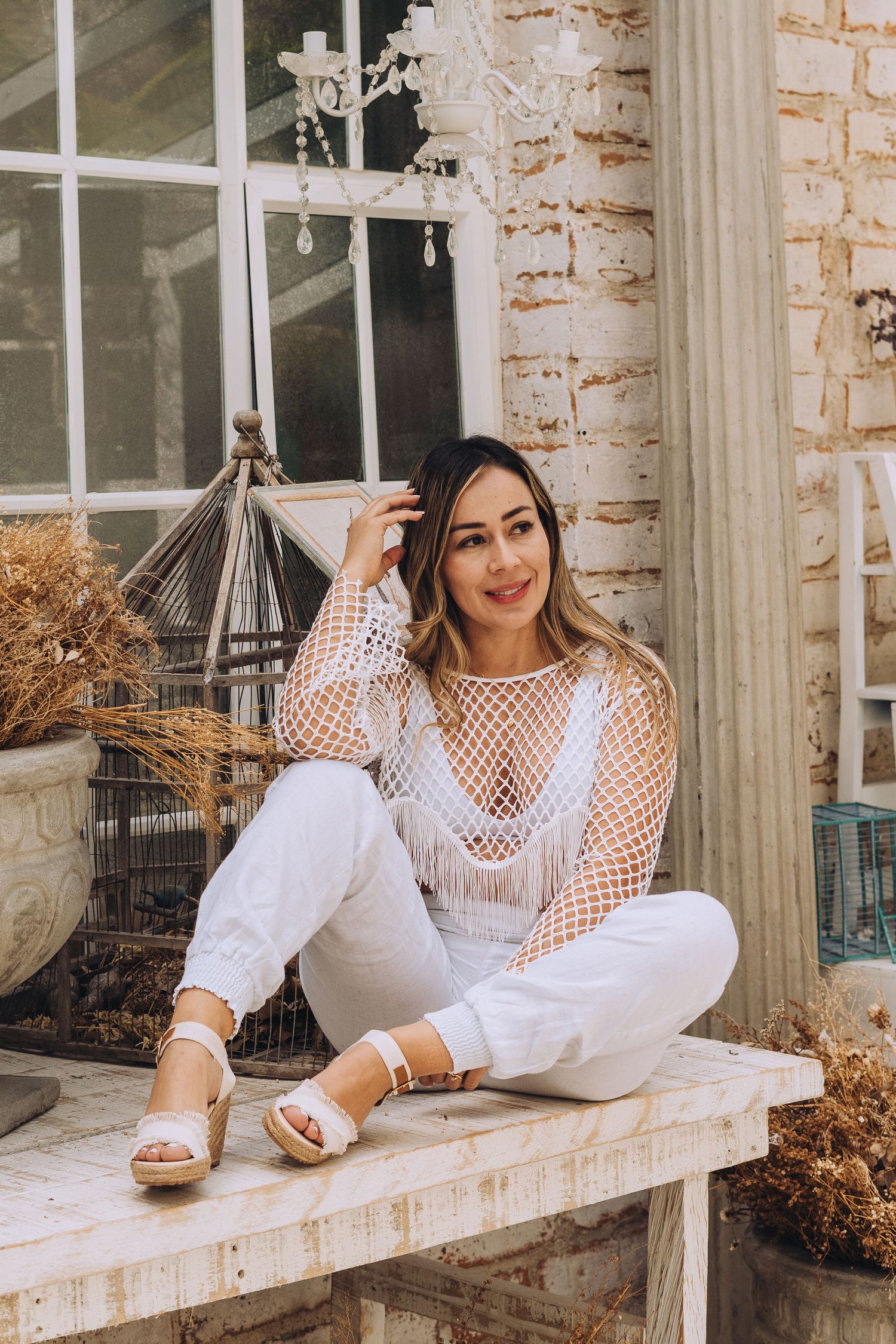Person in white mesh top sitting on rustic bench by vintage window and birdcage decor.