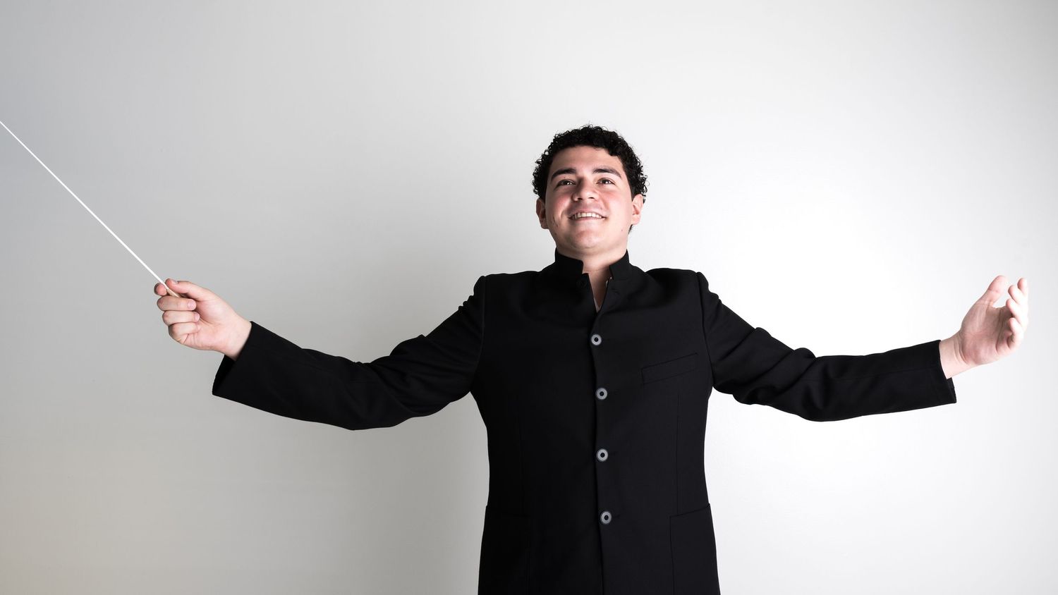 Orchestra conductor in black suit with arms spread wide holding batons against white background.
