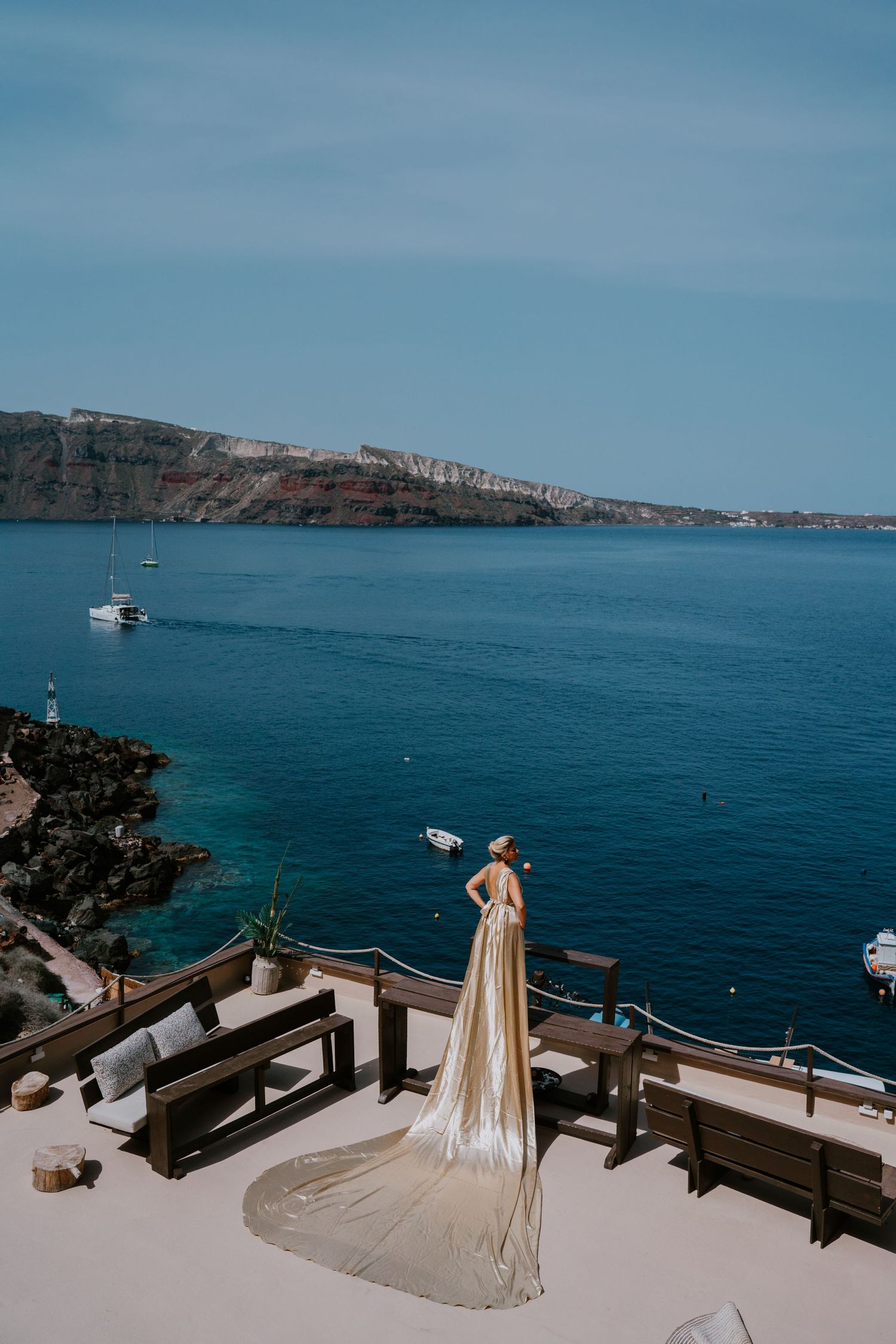 Long cream colored dress trails elegantly on a white terrace overlooking Mediterranean waters and cliffs.