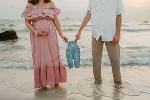 Expectant couple holding tiny denim overalls on beach at sunset.