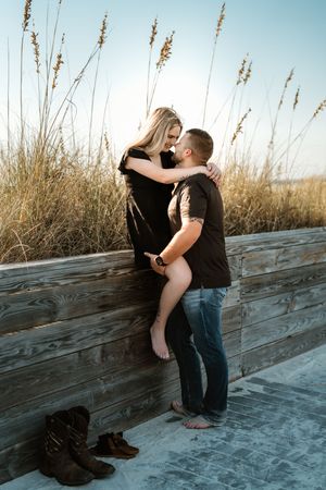 A romantic couple embraces on a wooden boardwalk near beach grass during sunset.