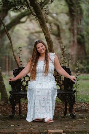 Person in white lace dress sits gracefully between two ornate black garden chairs in a dreamy woodland setting.