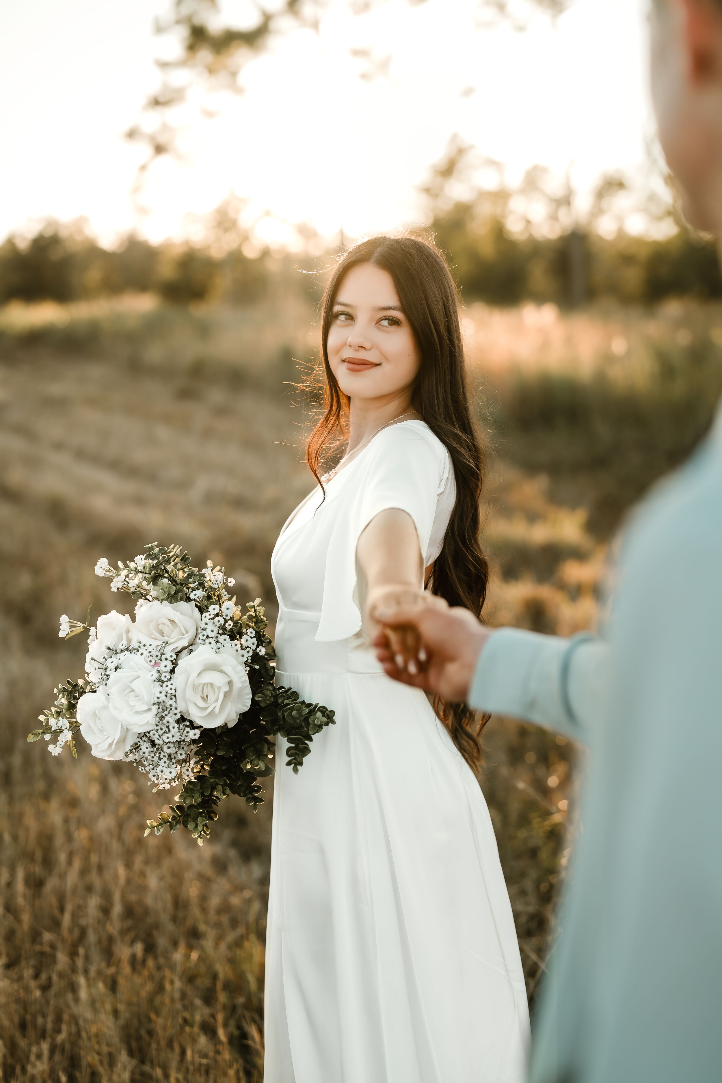 A person in a flowing white dress holds a delicate bouquet of white flowers during a golden hour photoshoot in a field.