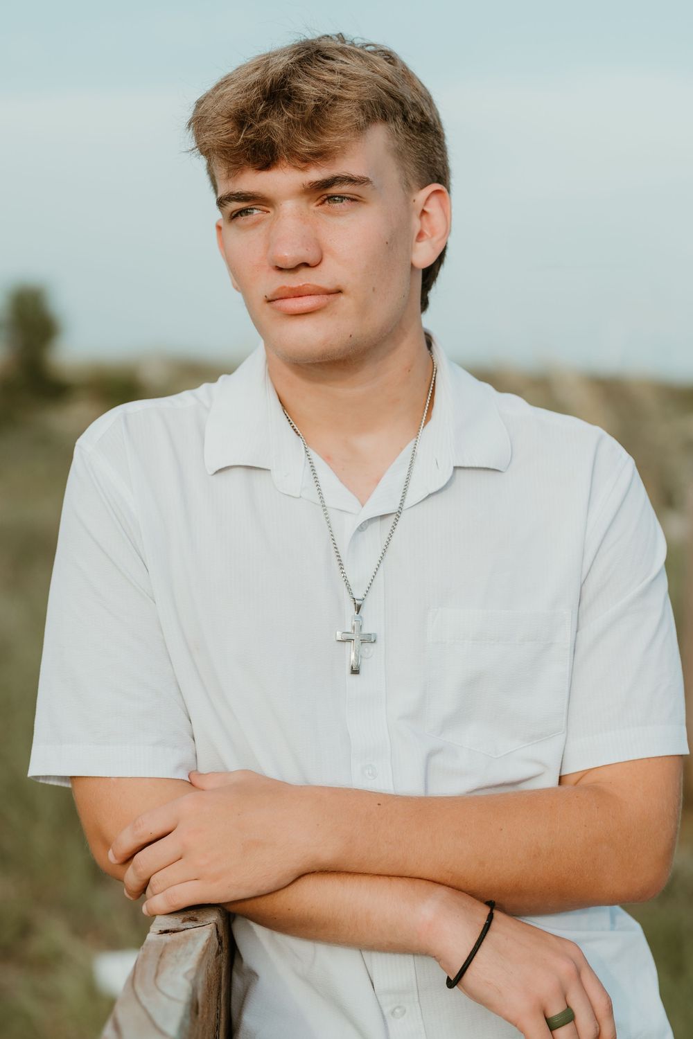 Young man in white shirt wearing cross necklace poses outdoors with arms crossed against scenic background.