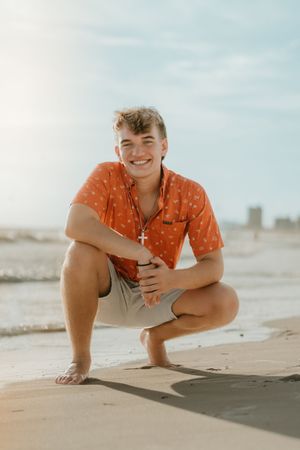 Young man in orange shirt crouching on beach, smiling, wearing cross necklace, with ocean waves in background.