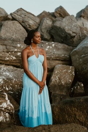 A person in a light blue tiered sundress poses against dark coastal rocks at sunset.