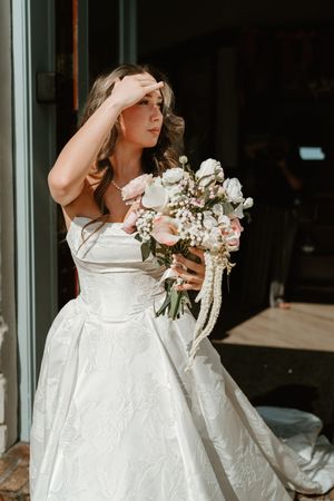 A bride in a white wedding dress holds a romantic bouquet of pink and white flowers against a window backdrop.