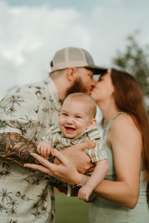 A happy couple kisses while holding their laughing baby outdoors, with the father wearing a floral shirt and trucker hat.