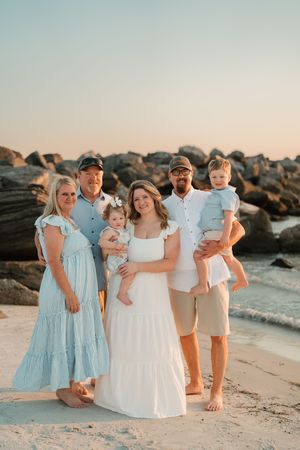 Family of six posing on a sandy beach at sunset, wearing coordinated blue and white outfits, with rocky shore in background.