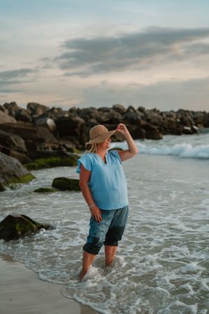 A woman in a blue top, denim shorts, and straw hat stands in ocean waves on a rocky beach at dusk.