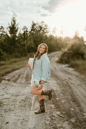 A person in a mint sweater and brown boots poses playfully on a sandy dirt path during golden hour.
