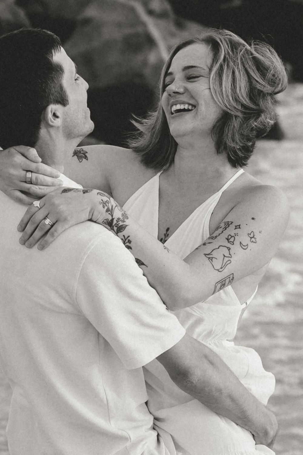 Black and white photo of a laughing couple embracing on a beach, both with tattoos, wearing white outfits.