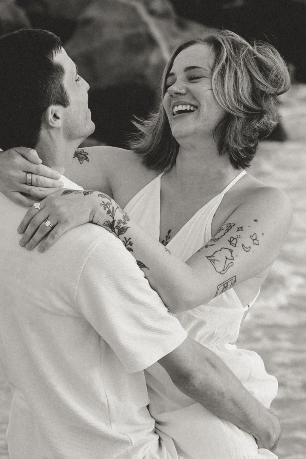 Black and white photo of a laughing couple embracing on a beach, both with tattoos, wearing white outfits.