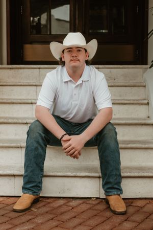 Someone wearing a white cowboy hat, white polo shirt and jeans sits on concrete steps in a western-style casual portrait.