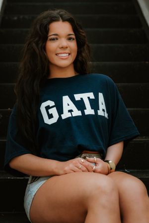 Someone sits on stairs wearing a navy GATA t-shirt and light colored shorts while smiling at the camera in warm lighting.