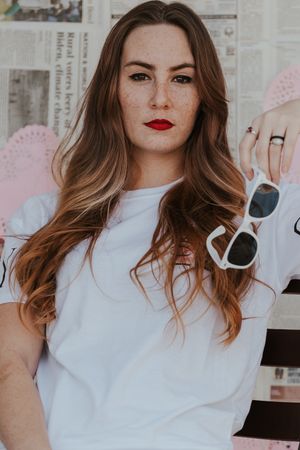 Lifestyle portrait of someone holding white sunglasses wearing red lipstick against newspaper background.