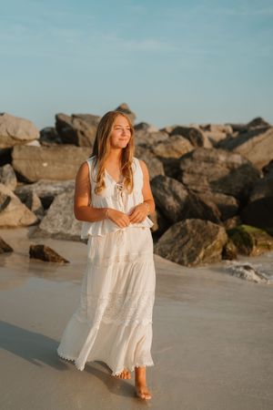 Young woman in white boho maxi dress walking barefoot on sandy beach with rocky backdrop at golden hour.