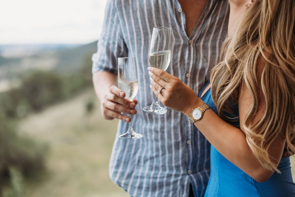 Couple clinking champagne glasses outdoors, celebrating with sparkling wine against a scenic green landscape backdrop.