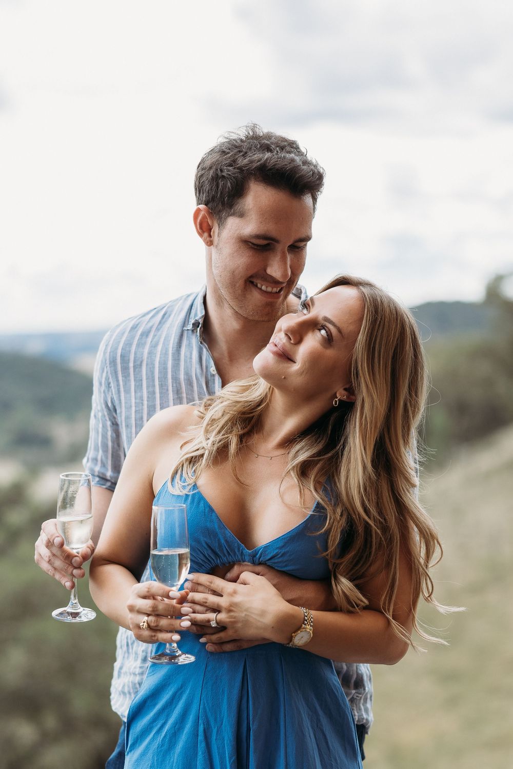A smiling couple embraces outdoors, holding champagne glasses, with a scenic green landscape in the background.
