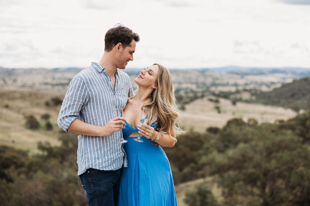 Couple embracing on a hilltop overlooking a scenic valley, woman in blue dress smiling at man in striped shirt.