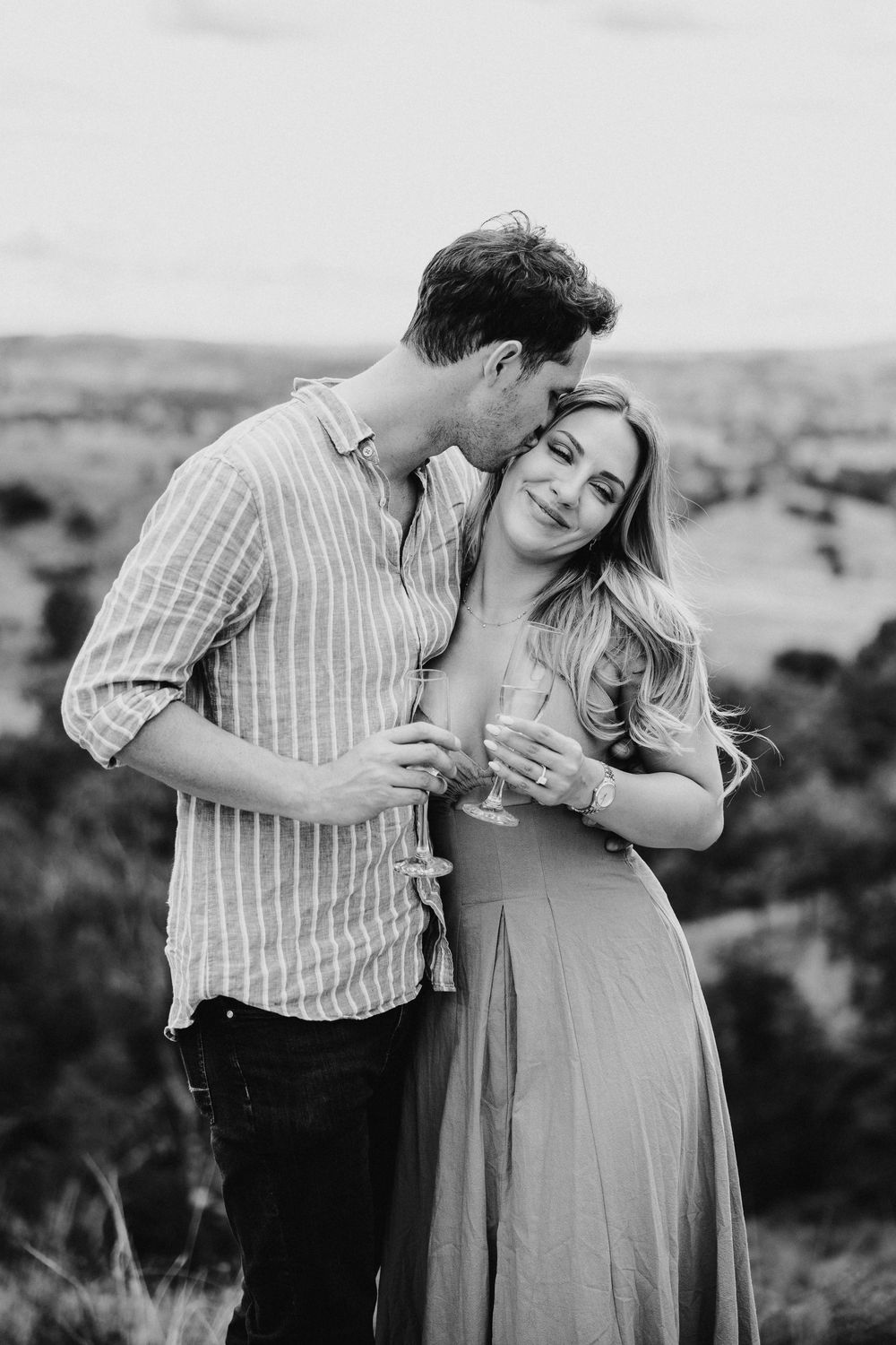 Black and white photo of a couple sharing a romantic moment outdoors, holding champagne glasses with scenic landscape behind.