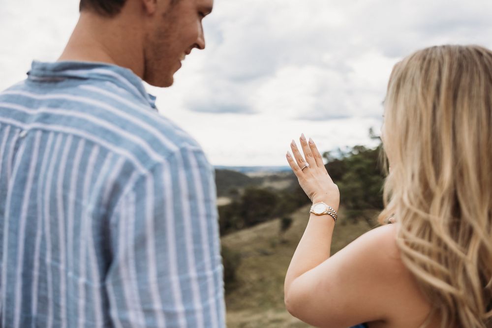 Couple outdoors admiring an engagement ring on woman's hand with scenic green hills in background.