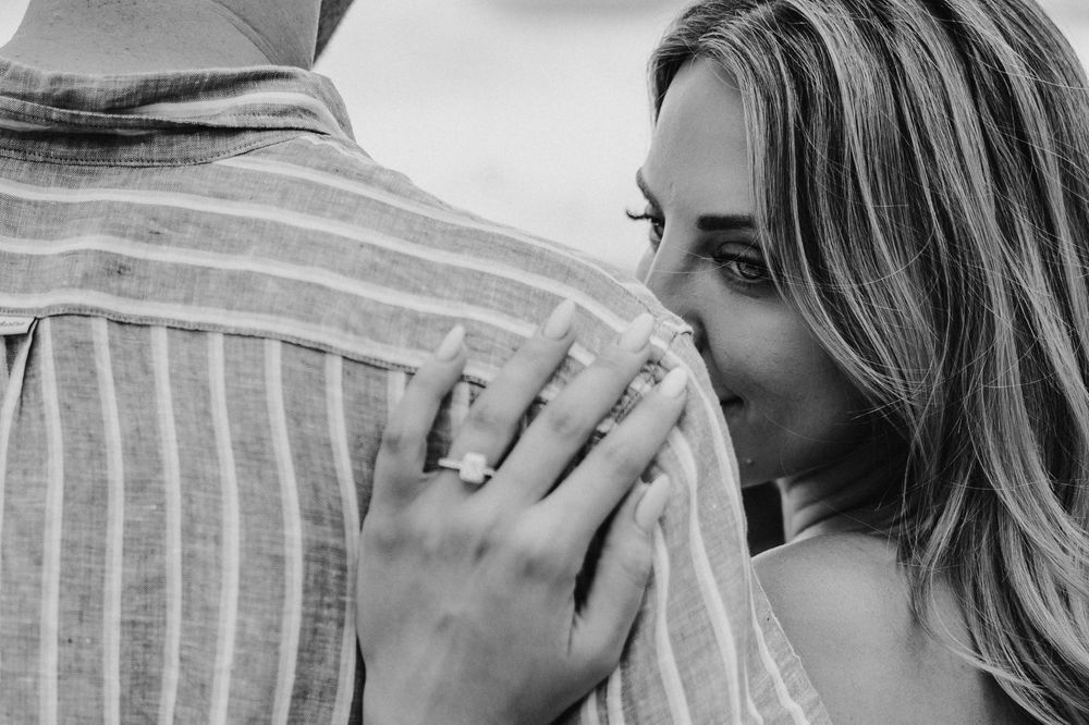 Black and white photo of a couple embracing, woman smiling with ring on finger, romantic engagement moment.