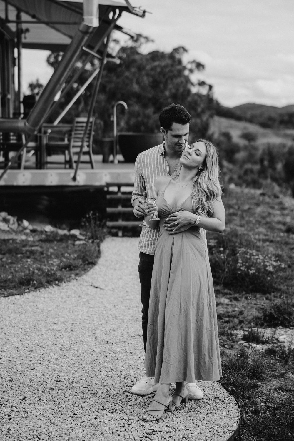 Black and white photo of a couple embracing on a gravel path outside a rural home, woman in maxi dress leaning back into mans arms.