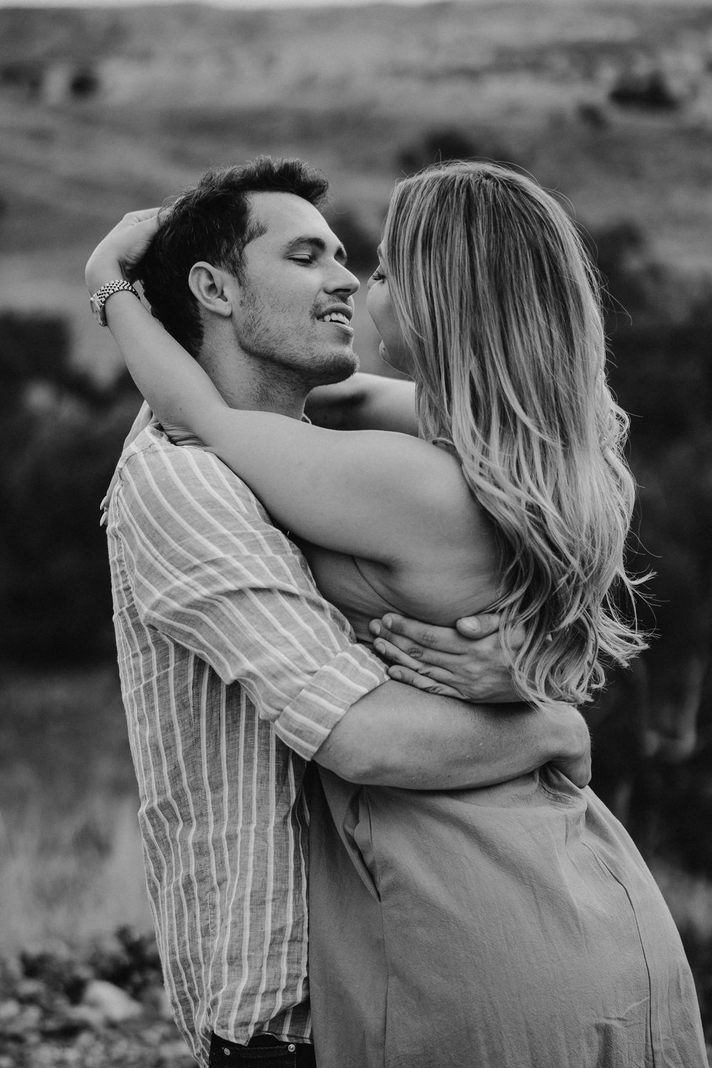 Black and white photo of a couple embracing outdoors, smiling at each other with rolling hills in the background.