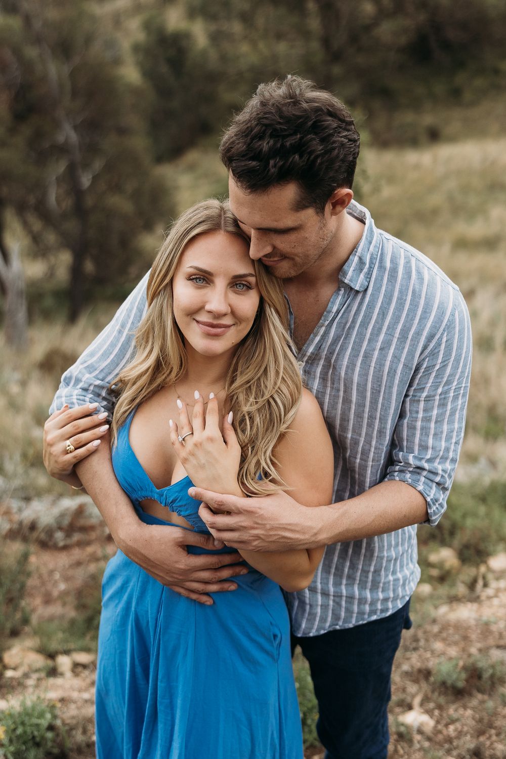 A couple embraces outdoors, woman in blue dress showing engagement ring, man in striped shirt kissing her head.