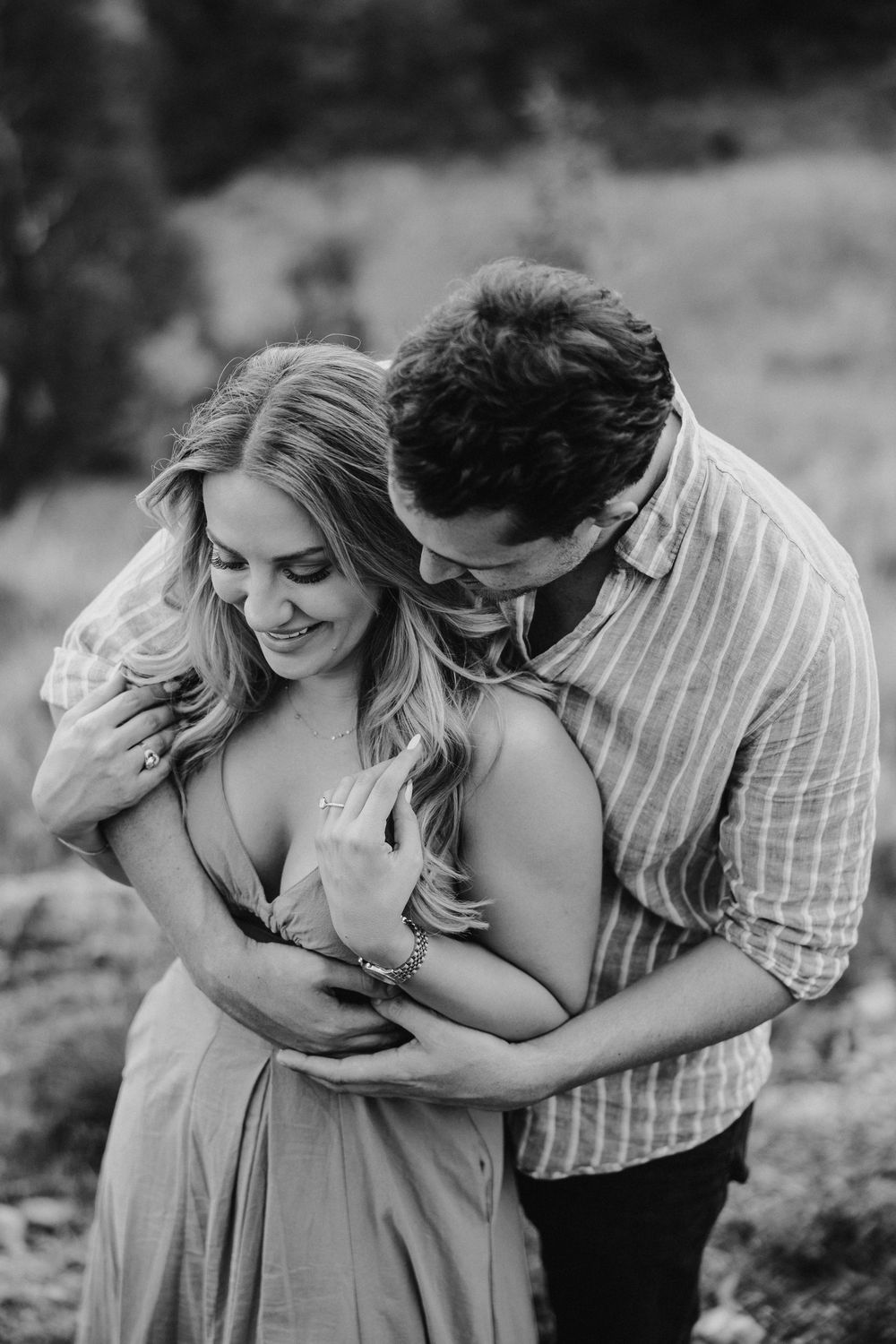 Black and white photo of a couple embracing outdoors, man holding woman from behind as she smiles warmly.