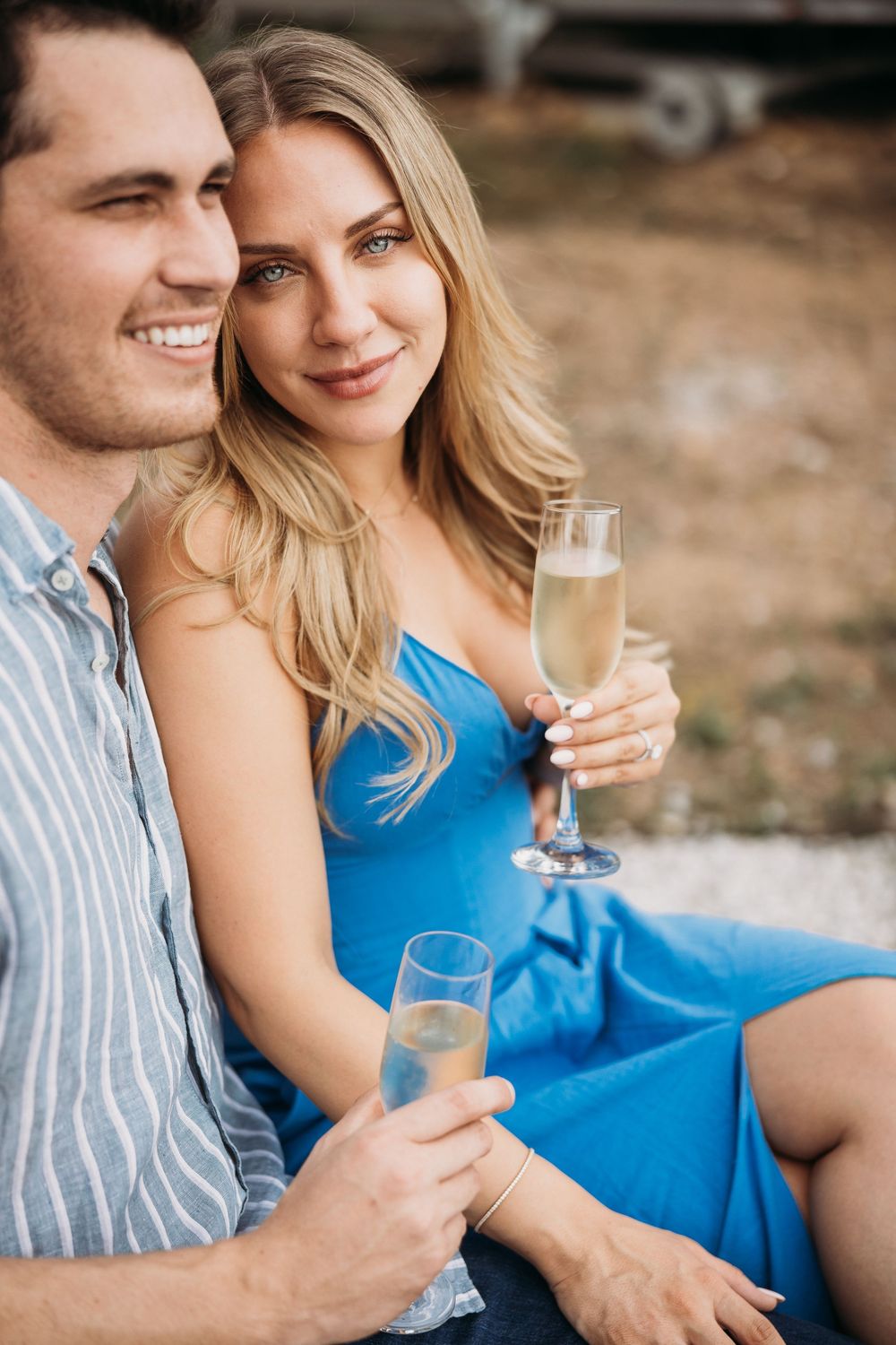 Happy couple sitting outdoors, clinking champagne glasses, woman in blue dress smiling at camera.