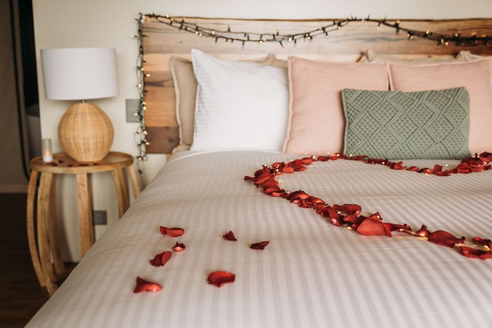 Romantic bedroom with rose petals arranged on white bedding, pink pillows, fairy lights, and a wooden nightstand with lamp.