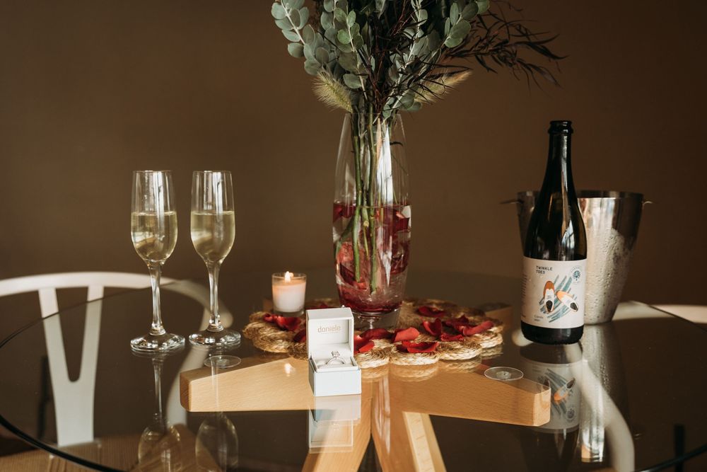 Romantic table setting with champagne flutes, rose petals, candle, desserts, and a wine bottle in a bucket.