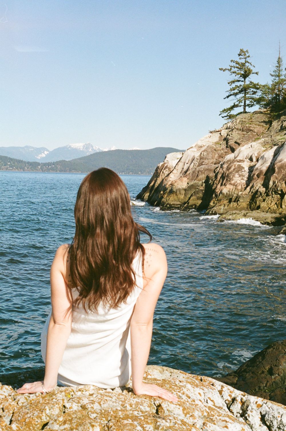 Person relaxes on rocky seaside cliff overlooking blue ocean waters and mountain vista.