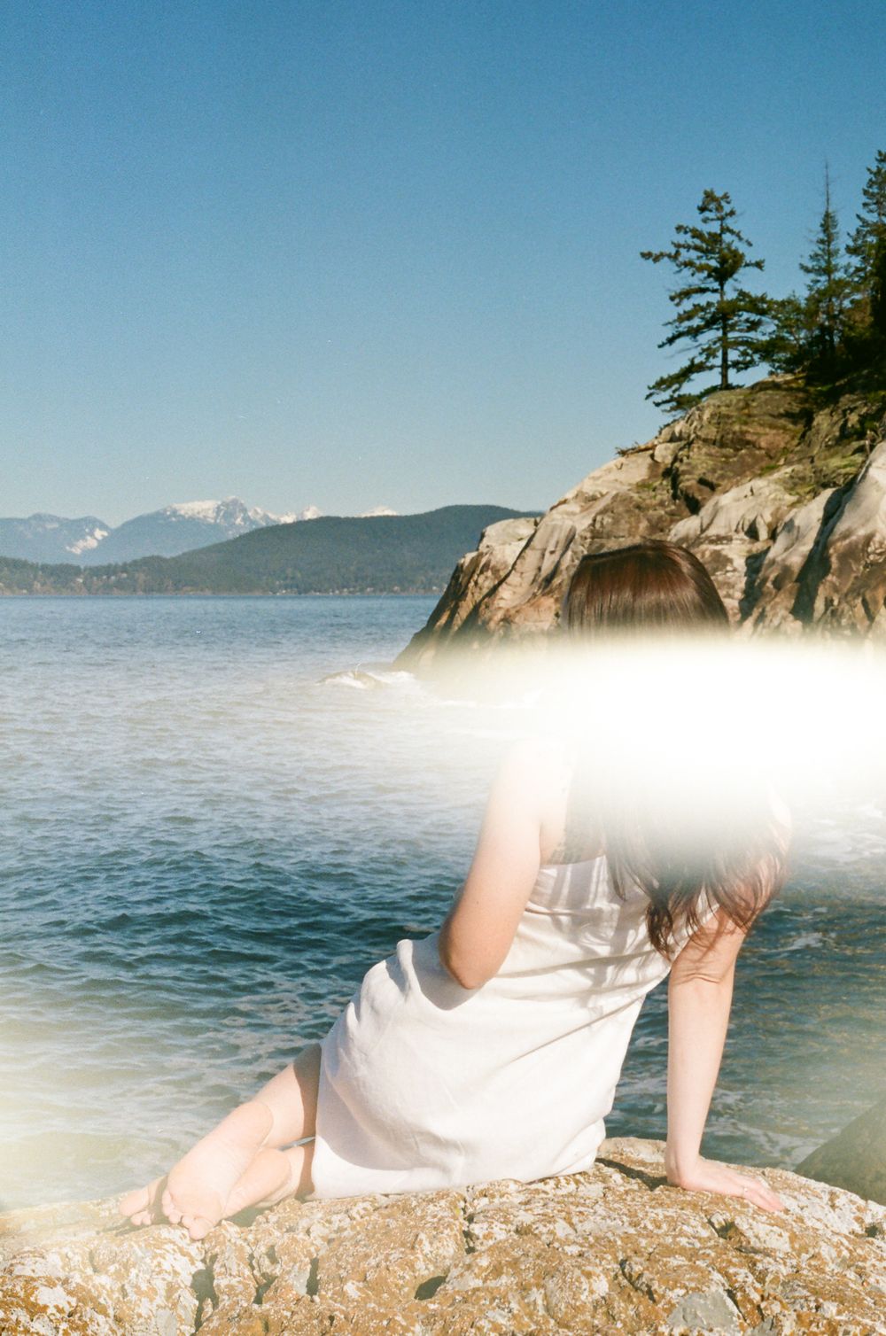 Person in white dress reclines on rocky coastal outcrop with mountains and ocean in background.
