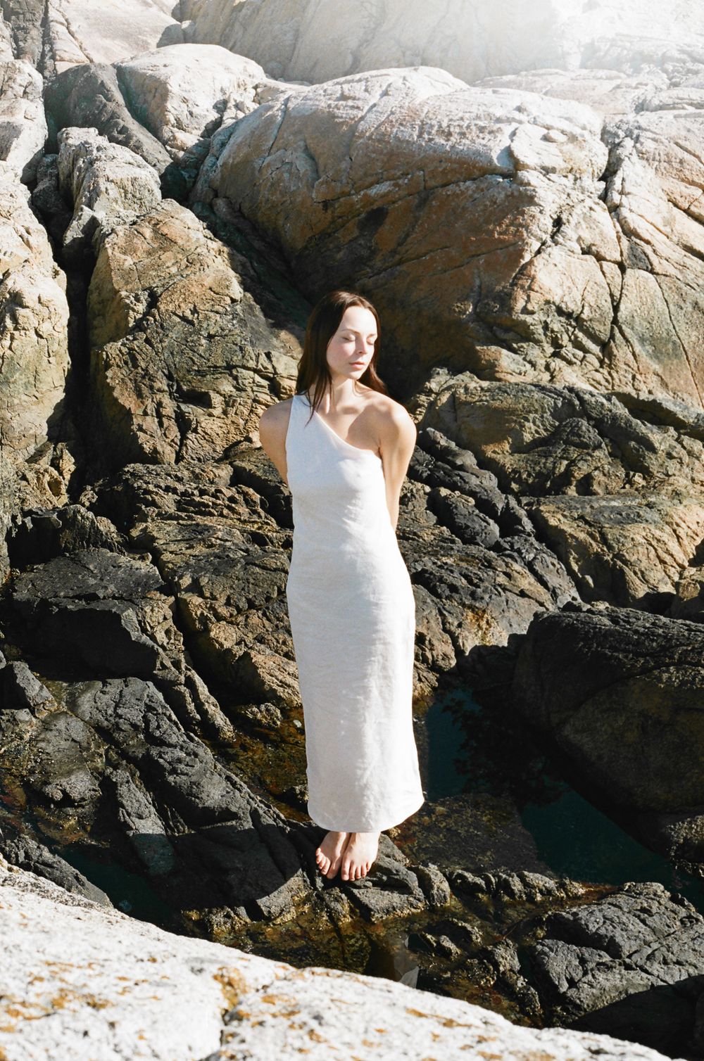 Figure in white dress poses against dramatic rocky coastal backdrop.