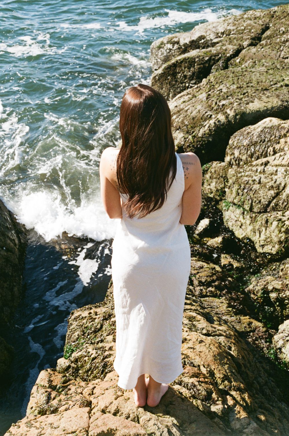 Person in white sits on rugged coastal rocks as waves crash against shore.