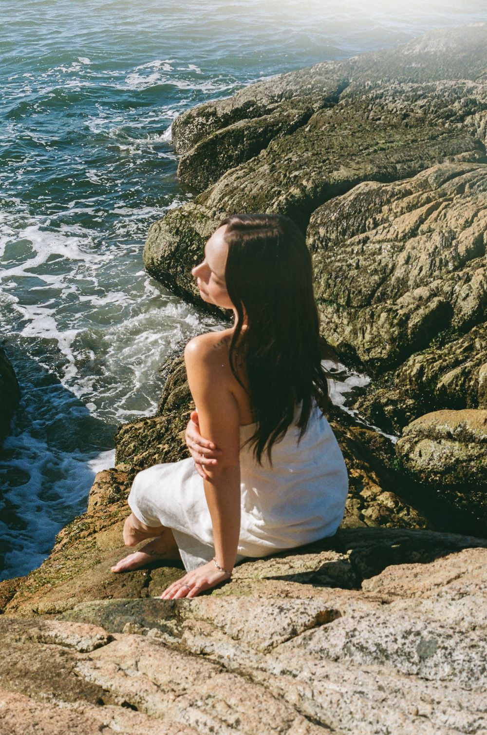 Figure rests on rocky cliff edge with crashing waves and ocean view.