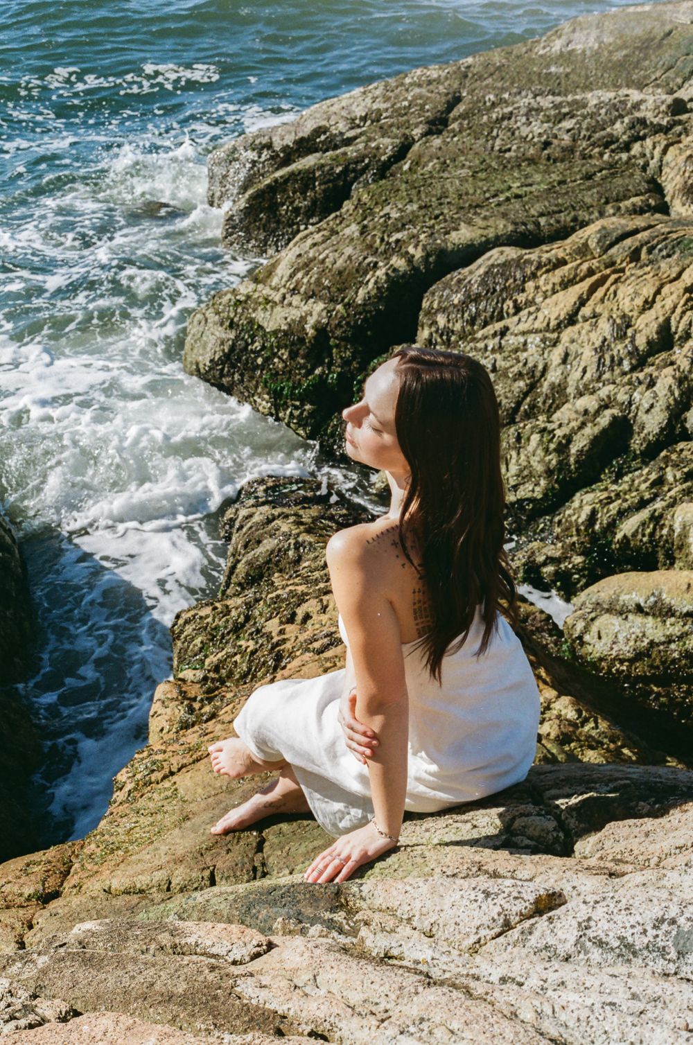 Figure in white dress reclines on rocky shoreline beside crashing ocean waves.