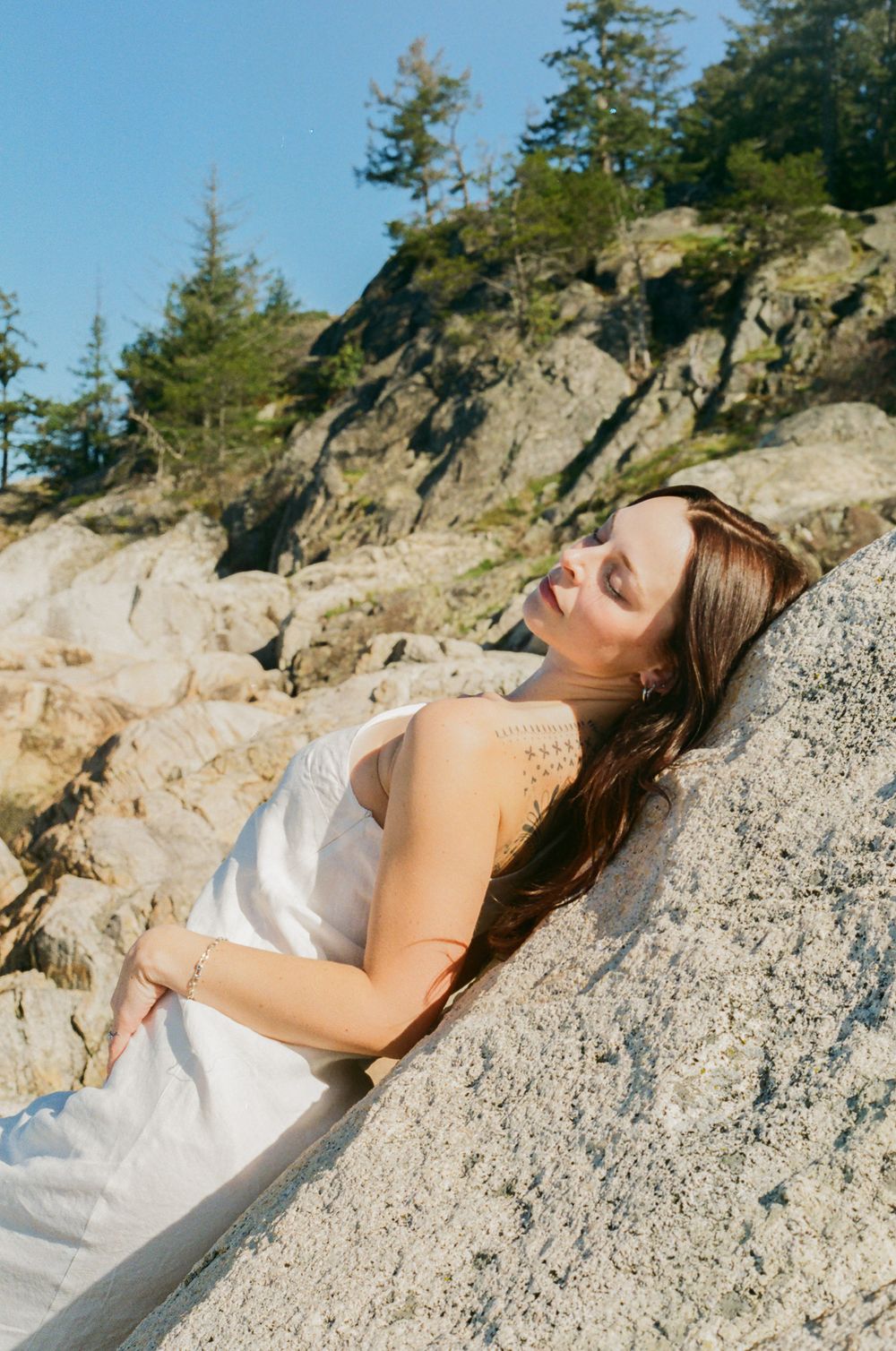 Person lounges on sun-drenched coastal rock formation with evergreen trees in background.
