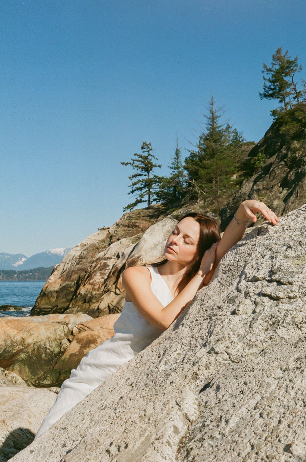 Person relaxes on sunlit coastal rocks with mountain vista and blue ocean in background.