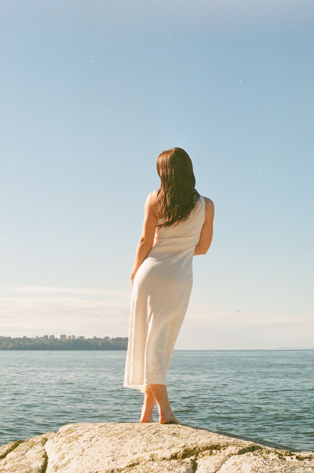 Figure in white dress stands on rocky outcrop overlooking calm ocean waters.