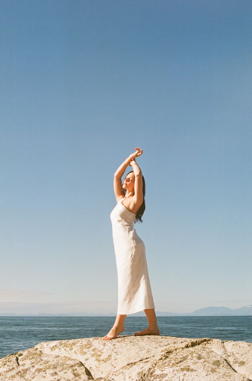 Person in flowing white dress poses gracefully on sunlit coastal rocks by the ocean.