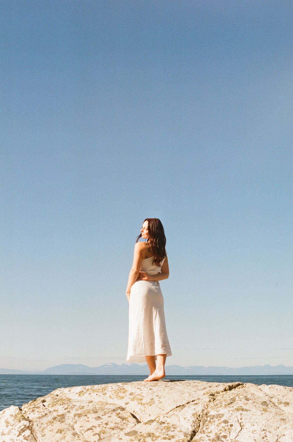 Figure in white dress poses on rocky coastline against blue ocean and sky background.