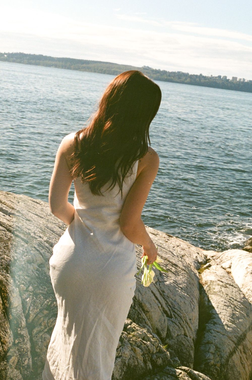 Person in white dress sits on rocky coastal outcrop overlooking ocean waters.