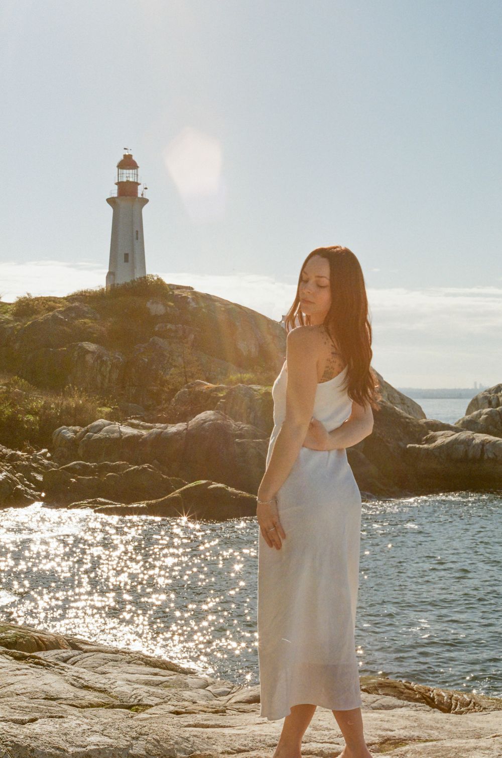 Lighthouse scene with figure in white dress standing on rocky shore during golden hour.